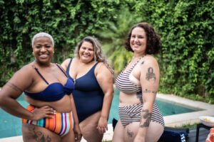 Three happy obese women standing in their swimsuits in front of an outdoor pool.